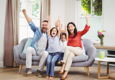 família com pai, mãe e duas filhas comemorando no sofá da sala