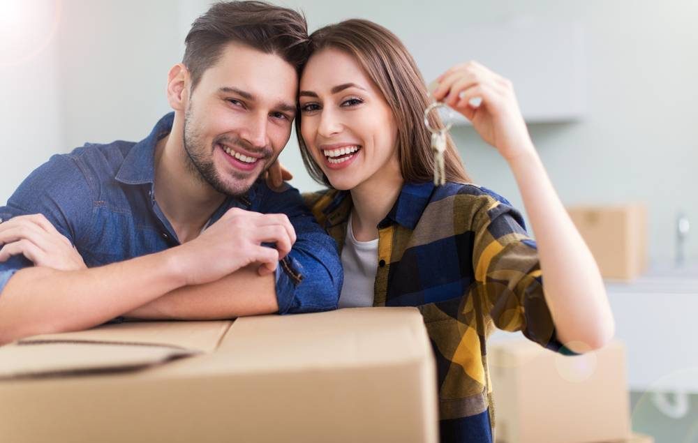 casal jovem sorrindo, segurando uma chave com os braços apoiados numa caixa de papelão.
