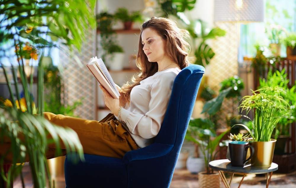 mulher jovem branca lendo livro em uma varanda repleta de plantas, sentada em uma poltrona azul