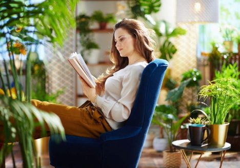 mulher jovem branca lendo livro em uma varanda repleta de plantas, sentada em uma poltrona azul