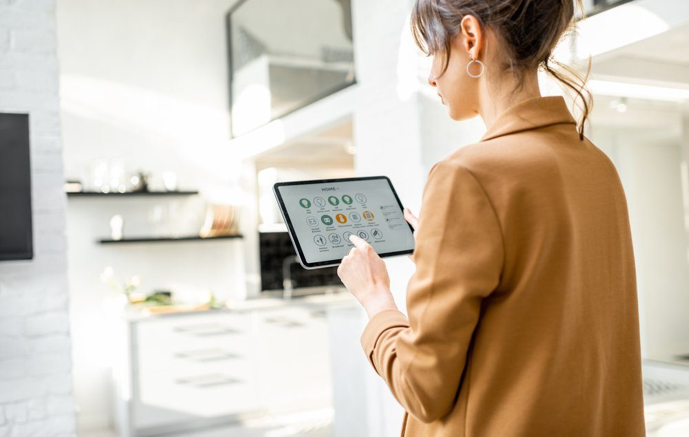 mulher branca de cabelo castanho escuro, vestindo um casaco caramelo, olhando para um tablet com aplicativos de smart home