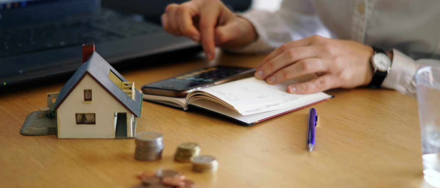 homem em mesa fazendo calculos e maquete de casa com moedas ao lado